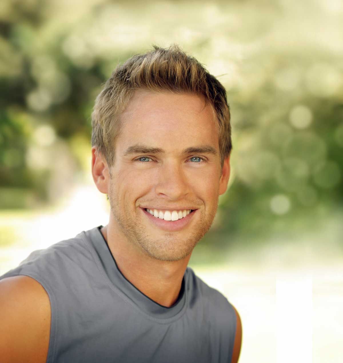Young man with bright white smiling outside against a blurred green background.